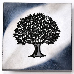May include: A black metal stamp featuring a tree design with a thick trunk and numerous leaves. The stamp is set against a silver background.