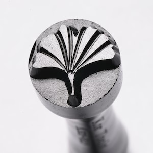 May include: A close-up of a metal stamp featuring a ginkgo leaf design. The stamp is round and has a textured surface.