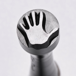 May include: A metal hand stamp with a raised design of a hand with five fingers. The stamp is round and has a black finish.