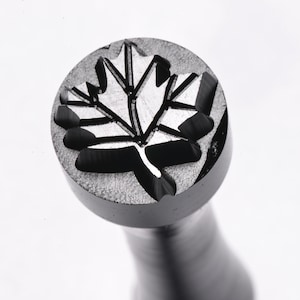 May include: A metal stamp with a detailed maple leaf design. The stamp is round and has a handle.