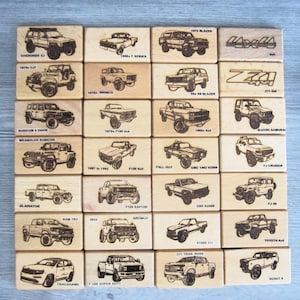 May include: A collection of rectangular wooden blocks, each featuring a laser-engraved illustration of a different truck or SUV model. The designs include various off-road vehicles, pickups, and classic cars, with the name of each vehicle printed below.