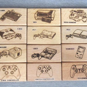 May include: A set of twelve rectangular wooden blocks, each featuring a laser-engraved illustration of a video game console or controller. The labels include Atari 2600, NES, Sega Genesis, X Box, PS 1, PS 3, Gamecube, SNES, Gameboy, Xbox controller, Switch controller, and PS 5 controller.