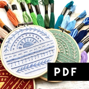 Puede incluir: Dos aros de bordado con diseños diferentes. El aro de la izquierda tiene un diseño azul y blanco con un sol y hojas. El aro de la derecha tiene un diseño verde y blanco con hojas y puntos. Los aros están rodeados de hilo de bordar de colores.