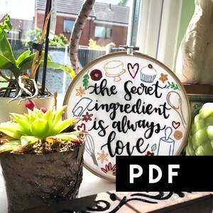 May include: Embroidered hoop with the text "the secret ingredient is always love" surrounded by various kitchen-themed embroidery designs, including a cake, a cupcake, a whisk, a rolling pin, and a spoon. The hoop is white with a wooden frame and is sitting on a windowsill.