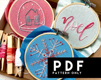 May include: Three embroidery hoops with different designs. The first hoop has a pink background with a white house inside a snow globe. The second hoop has a light blue background with the word "Noel" embroidered in pink. The third hoop has a blue background with a white snowflake embroidered in red and white.