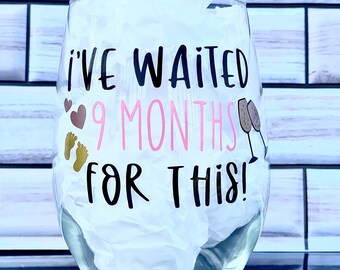 May include: A clear glass wine glass with a black and pink vinyl decal that reads "I've waited 9 months for this!" with a pink heart, two gold footprints, and two gold champagne glasses.