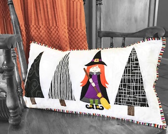 Puede incluir: Un cojín rectangular con temática de Halloween que presenta una bruja y tres árboles estilizados. La bruja tiene pelo rojo, un sombrero negro y un vestido morado. Los árboles tienen patrones en blanco y negro. El cojín tiene un borde a rayas.