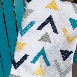 May include: A white quilt with a geometric pattern of triangles in shades of teal, navy, gray, and yellow. The quilt is made with different patterned fabrics, including polka dots, zigzags, and gingham.