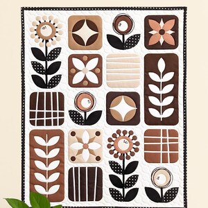 May include: A decorative quilt featuring a repeating pattern of stylized flowers and geometric shapes in brown, beige, and black on a white background. The quilt has a black border. A potted plant and decorative objects are on a wooden table.
