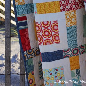 May include: A colorful patchwork quilt with a variety of patterns and colors, including red, blue, yellow, and white. The quilt is made up of squares and rectangles, and the patterns include flowers, waves, and geometric shapes. The quilt is hanging on a wooden fence.