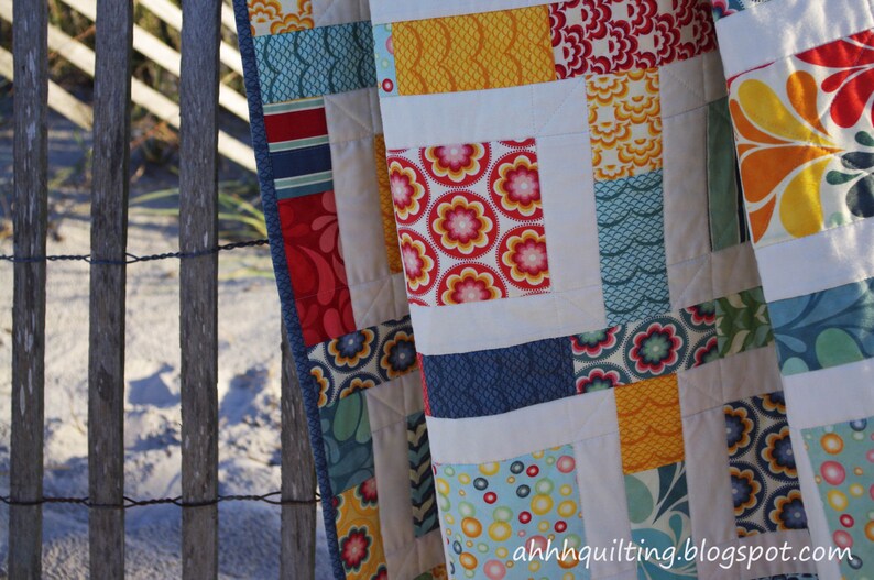 May include: A colorful patchwork quilt with a variety of patterns and colors, including red, blue, yellow, and white. The quilt is made up of squares and rectangles, and the patterns include flowers, waves, and geometric shapes. The quilt is hanging on a wooden fence.