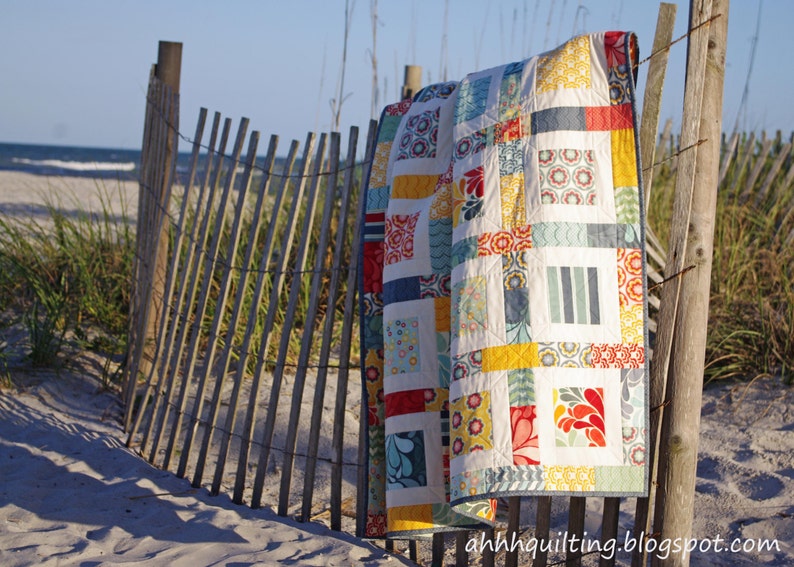 May include: A colorful patchwork quilt with a variety of geometric patterns and bright colors, including red, yellow, blue, and green. The quilt is hanging on a wooden fence against a sandy beach background.