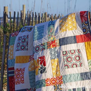 May include: A colorful patchwork quilt with a variety of floral and geometric patterns in shades of blue, red, yellow, and white. The quilt is hanging on a wooden fence against a backdrop of a beach and ocean.