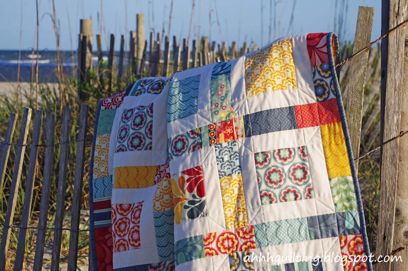 May include: A colorful patchwork quilt with a variety of floral and geometric patterns in shades of blue, red, yellow, and white. The quilt is hanging on a wooden fence against a backdrop of a beach and ocean.