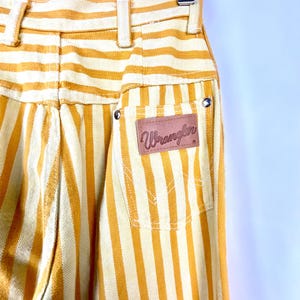 K&ouml;nnte beinhalten: Nahaufnahme einer Vintage-Wrangler-Streifenhose in Gelb und Creme. Die Hose hat vertikale Streifen, einen braunen Lederaufn&auml;her mit dem Wrangler-Logo und klassische Taschen-Details. Die Hose ist hoch tailliert und hat eine klassische Passform.