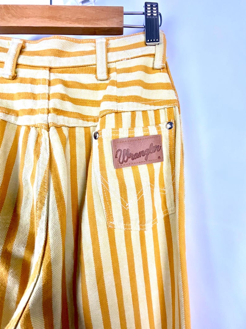 K&ouml;nnte beinhalten: Nahaufnahme einer Vintage-Wrangler-Streifenhose in Gelb und Creme. Die Hose hat vertikale Streifen, einen braunen Lederaufn&auml;her mit dem Wrangler-Logo und klassische Taschen-Details. Die Hose ist hoch tailliert und hat eine klassische Passform.
