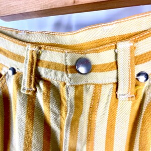 K&ouml;nnte beinhalten: Nahaufnahme einer gelb-wei&szlig; gestreiften Hose. Die Hose hat einen Knopfverschluss und G&uuml;rtelschlaufen. Der Stoff scheint eine gewebte Baumwoll- oder Leinenmischung zu sein. Die Hose h&auml;ngt an einem Holzb&uuml;gel.