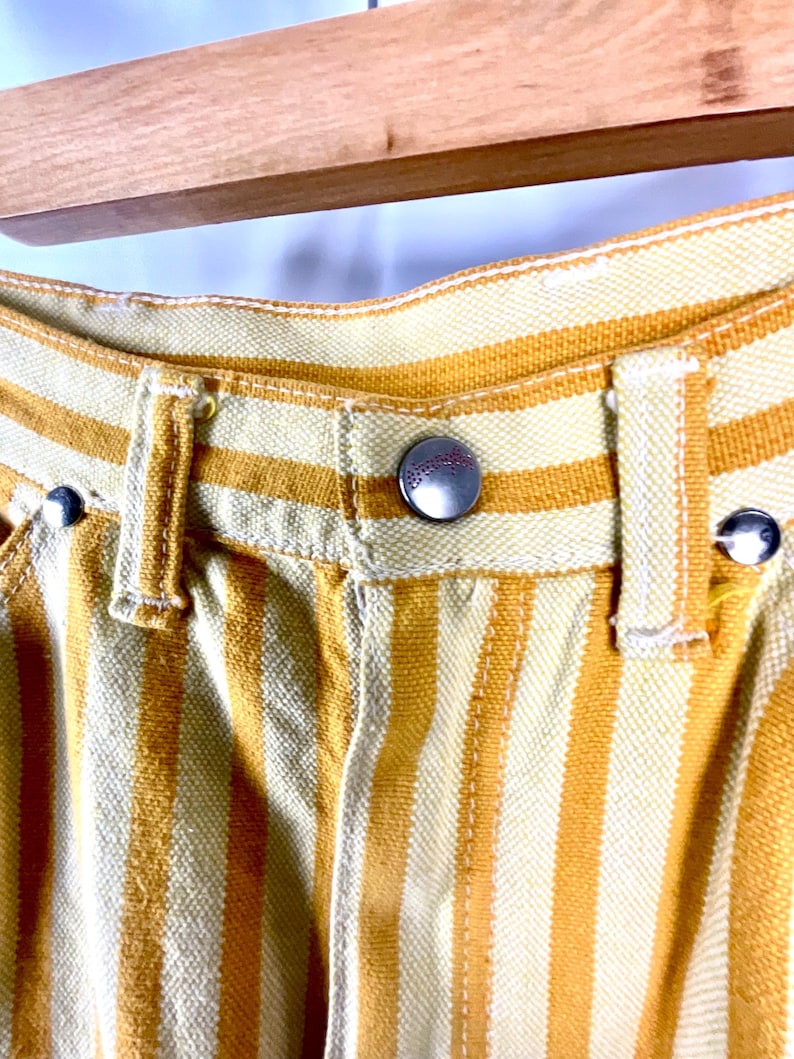 K&ouml;nnte beinhalten: Nahaufnahme einer gelb-wei&szlig; gestreiften Hose. Die Hose hat einen Knopfverschluss und G&uuml;rtelschlaufen. Der Stoff scheint eine gewebte Baumwoll- oder Leinenmischung zu sein. Die Hose h&auml;ngt an einem Holzb&uuml;gel.