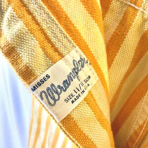 K&ouml;nnte beinhalten: Nahaufnahme eines Vintage-Kleidungsst&uuml;cks mit vertikalen Streifen in Gelb- und Oranget&ouml;nen. Ein Etikett lautet "Misses Wrangler" mit der Gr&ouml;&szlig;e 11/11 Slim und "Made in U.S.A."