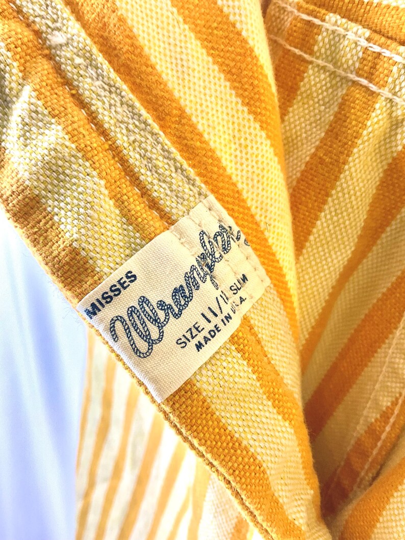 K&ouml;nnte beinhalten: Nahaufnahme eines Vintage-Kleidungsst&uuml;cks mit vertikalen Streifen in Gelb- und Oranget&ouml;nen. Ein Etikett lautet "Misses Wrangler" mit der Gr&ouml;&szlig;e 11/11 Slim und "Made in U.S.A."