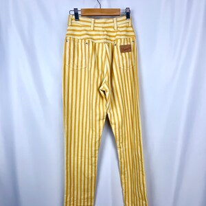 K&ouml;nnte beinhalten: Hochtaillierte Hose mit gelb-wei&szlig;em Streifenmuster, h&auml;ngend an einem Holzb&uuml;gel. Die Hose hat ein klassisches F&uuml;nf-Taschen-Design und einen braunen Lederaufn&auml;her. Die vertikalen Streifen erzeugen einen Schlankheitseffekt.