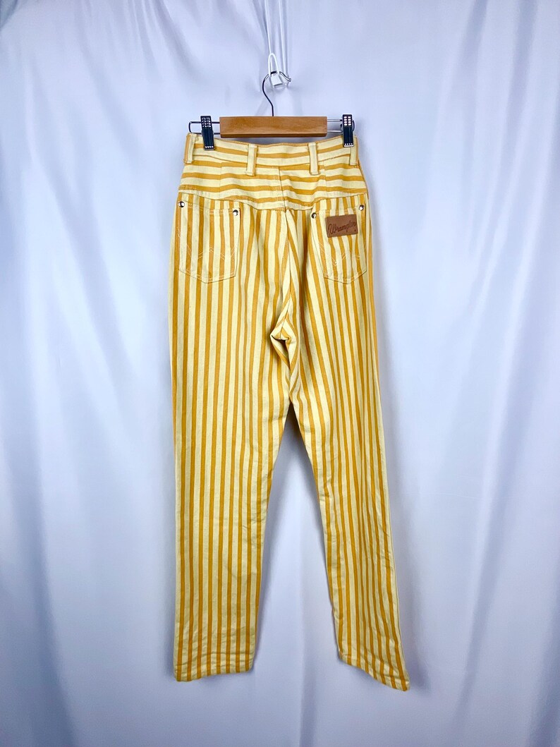 K&ouml;nnte beinhalten: Hochtaillierte Hose mit gelb-wei&szlig;em Streifenmuster, h&auml;ngend an einem Holzb&uuml;gel. Die Hose hat ein klassisches F&uuml;nf-Taschen-Design und einen braunen Lederaufn&auml;her. Die vertikalen Streifen erzeugen einen Schlankheitseffekt.
