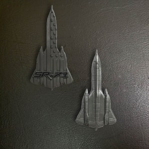 Op de afbeelding: Twee zilveren metalen modelvliegtuigen, een is een grotere SR-71 Blackbird met de tekst "SR-71" op de romp, de andere is een kleiner model van een vergelijkbaar vliegtuig.