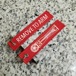 May include: Two red fabric keychains with white embroidery. One keychain says "REMOVE TO ARM" and the other has a star and a rocket ship design.