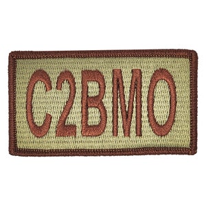 May include: Embroidered patch with the letters "C2BMO" in a reddish-brown color. The letters are stitched onto a light olive-green background, framed by a dark brown border. The patch is rectangular.
