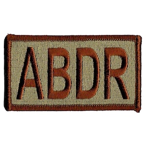 Può includere: Una toppa ricamata di colore beige e marrone con le lettere "ABDR" in marrone.