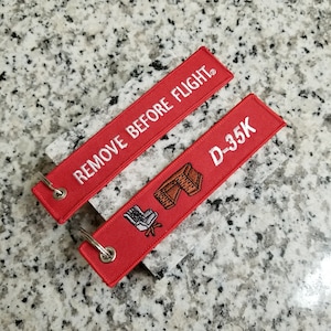 May include: Two red fabric keychains with white embroidery. One keychain says "REMOVE BEFORE FLIGHT" and the other says "D-35K" with a small embroidered image of a chair and a desk.