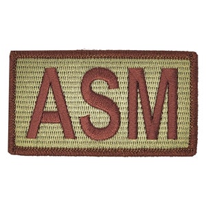 Puede incluir: Parche bordado rectangular con las letras "ASM" en color rojo oscuro. Las letras están cosidas sobre un fondo beige claro, enmarcado por un borde marrón. El parche presenta un aspecto texturizado.