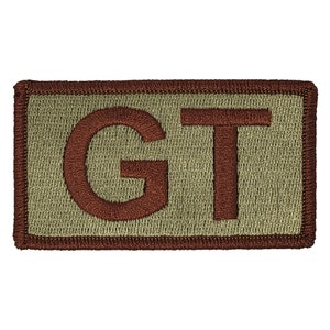 May include: A brown embroidered patch with the letters "GT" in a rectangular shape on a tan background.