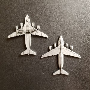 May include: Two silver metal model airplanes, both are C-17s. The top plane has a US flag on the fuselage and the text "C-17" on the nose. The bottom plane is a more detailed model with a raised tail.