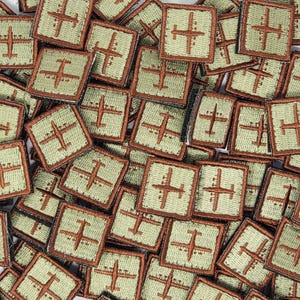 Op de afbeelding: Een close-up afbeelding van een stapel kleine, vierkante patches. Elke patch is groen met een bruine rand en heeft een geborduurd beeld van een vliegtuig. De patches zijn in een willekeurig patroon gerangschikt.