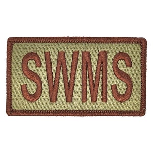 May include: Embroidered patch with the letters "SWMS" in a brown, textured font. The letters are on a light green background, framed by a brown border. The patch has a rectangular shape.