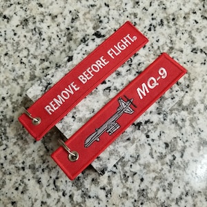 May include: Red fabric keychain with white text that says "REMOVE BEFORE FLIGHT" and a gray embroidered image of a drone with the text "MQ-9".