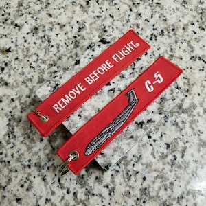 May include: Red and white fabric keychain with the text "REMOVE BEFORE FLIGHT" and a gray outline of a C-5 aircraft with the text "C-5".