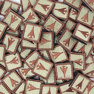 May include: A close-up image of a pile of brown and green embroidered patches featuring a brown airplane silhouette. The patches are square and have a brown border.