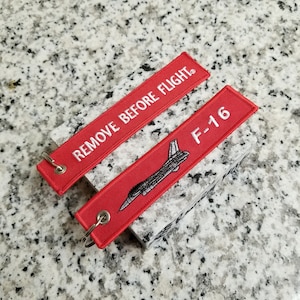 May include: Red keychain with a white airplane silhouette and the text "F-16". A second red keychain with the text "REMOVE BEFORE FLIGHT" is also shown.