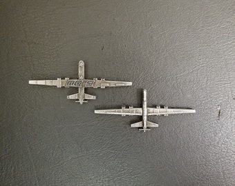 Könnte beinhalten: Zwei silberne Metallstifte in Form der MQ-9 Reaper Drohne. Der obere Stift trägt den Text "MQ-9".
