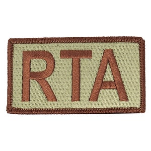 Puede incluir: Un parche rectangular de color beige con un borde bordado marrón y las letras "RTA" en marrón.