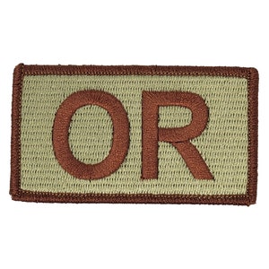 Peut inclure: Un patch rectangulaire de couleur beige avec une bordure brodée marron. Les lettres "OR" sont brodées en marron sur le patch.