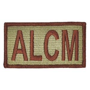 May include: A tan rectangular patch with a brown embroidered border and the letters "ALCM" in brown.