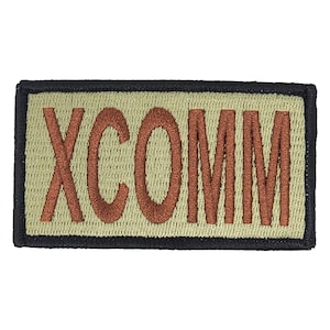 Op de afbeelding: Een rechthoekige, beige patch met een zwarte rand. Het woord "XCOMM" is geborduurd in bruin garen.