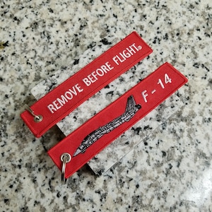 May include: Red fabric keychains with white text. One keychain says "REMOVE BEFORE FLIGHT" and the other says "F-14" with a black and white silhouette of a fighter jet.