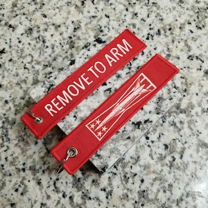 May include: Two red fabric keychains with white embroidery. One keychain says "REMOVE TO ARM" and the other has a white airplane design with three stars.