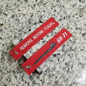 May include: Two red rectangular keychains with white text and silver metal rings. One keychain reads "REMOVE BEFORE FLIGHT." The other keychain has an illustration of an SR-71 aircraft and the text "SR-71."