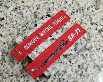 May include: Two red rectangular keychains with white text and silver metal rings. One keychain reads "REMOVE BEFORE FLIGHT." The other keychain has an illustration of an SR-71 aircraft and the text "SR-71."