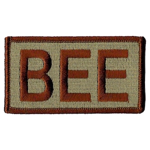 May include: A brown embroidered patch with the word "BEE" in brown letters on a tan background.